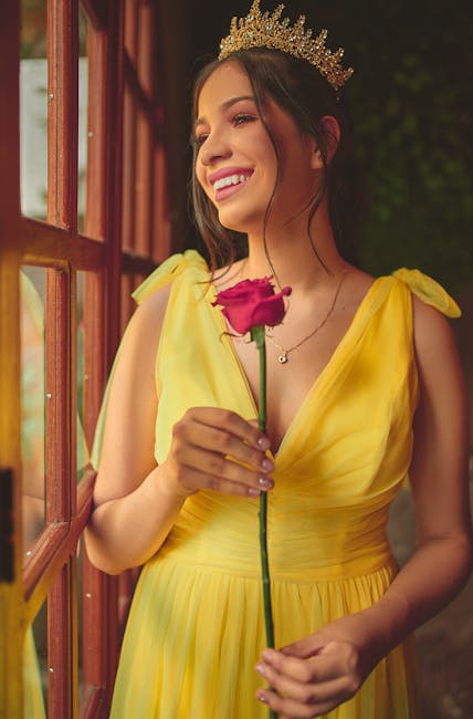 A woman with long dark hair wearing a golden crown and a bright yellow dress with puffed sleeves, standing near a wooden window frame in a garden setting. She is smiling softly, holding a single pink 