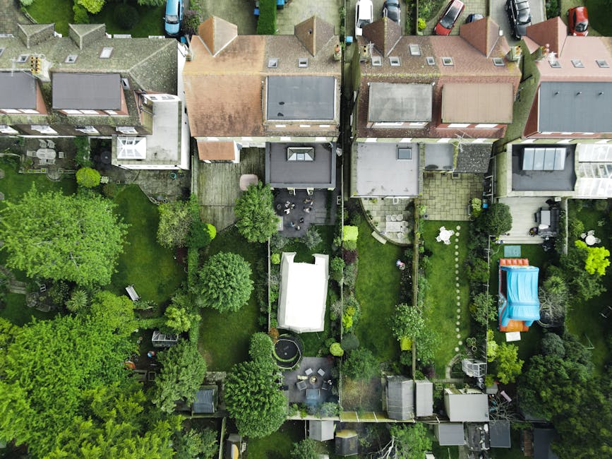 An aerial view of a small urban garden attached to a terraced house, showcasing a well-maintained green lawn bordered by lush trees and shrubs with dense foliage. The garden features a paved patio are